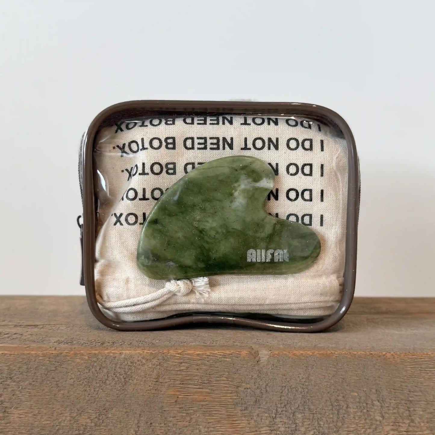 Green jade stone gua sha face massaging tool in a decorative plastic pouch with text on a wooden surface