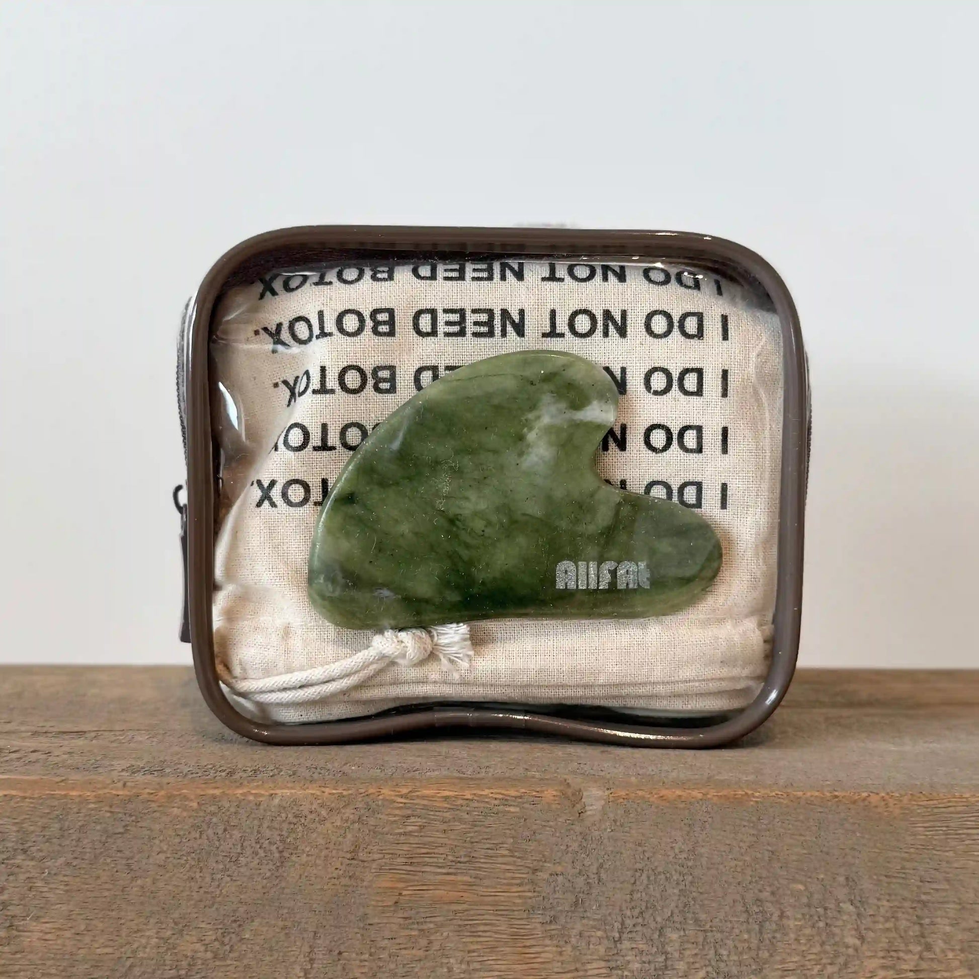 Green jade stone gua sha face massaging tool in a decorative plastic pouch with text on a wooden surface