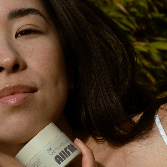 Woman holding 50 ml glass jar of ALLFAT Om Calming Tallow Cream while laying on a backdrop of natural leaves. 