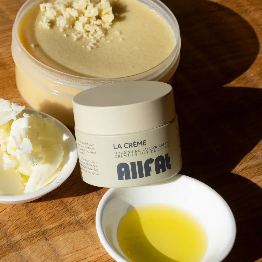 Jar of 'La Crème' tallow cream with a scoop taken out, surrounded by a bowl of cream and a small dish of oil on a wooden surface.