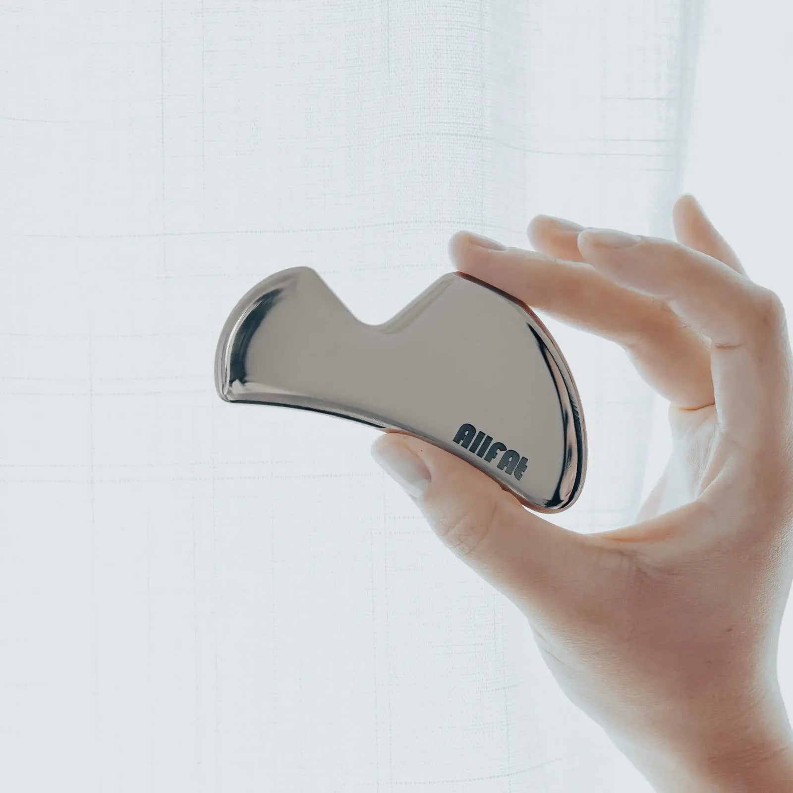 Hand holding a stainless steel gua sha face massaging tool with 'allfat' branding against a light background